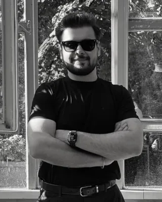 Osman Yağmur in a black T-shirt, arms folded, standing in front of open windows. Black and white photograph.